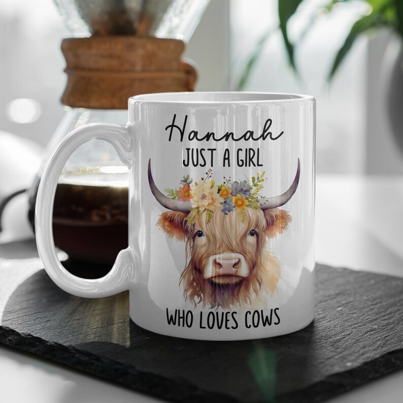 May include: White ceramic mug featuring a watercolor illustration of a Highland cow with floral crown. The mug has the text "Hannah JUST A GIRL WHO LOVES COWS" in black script. The mug is on a black slate surface.