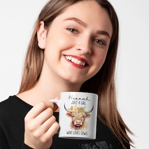 May include: White ceramic mug featuring a watercolor illustration of a Highland cow with floral accents and the text "Hannah JUST A GIRL WHO LOVES COWS". The mug has a curved handle.