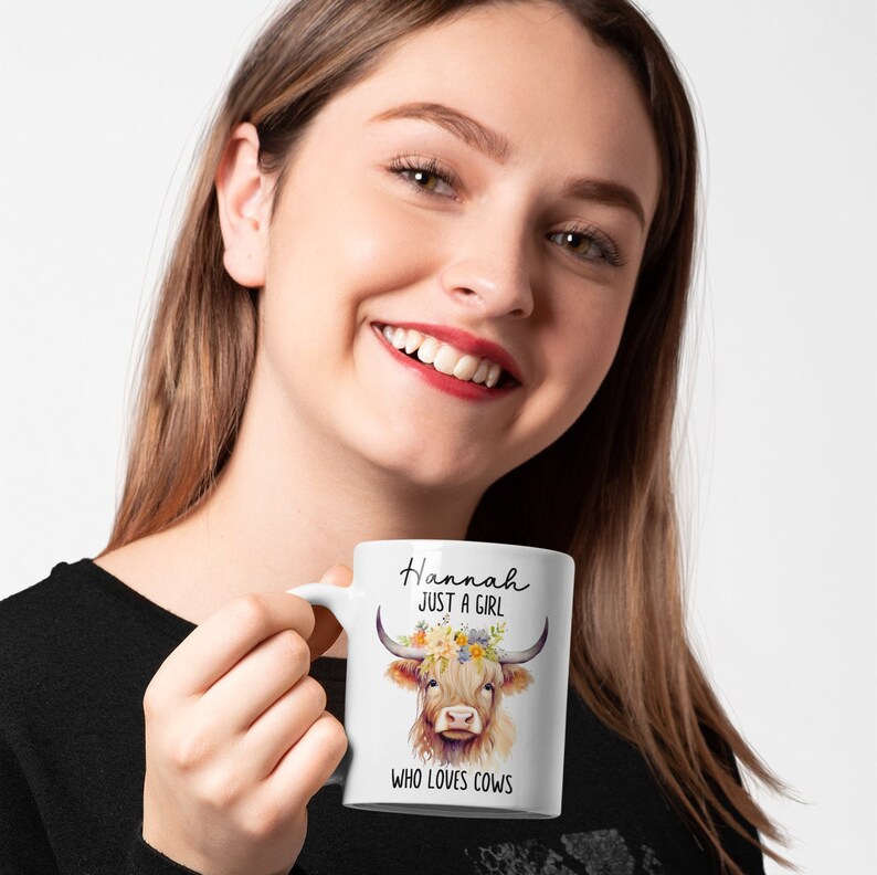 May include: White ceramic mug featuring a watercolor illustration of a Highland cow with floral accents and the text "Hannah JUST A GIRL WHO LOVES COWS". The mug has a curved handle.