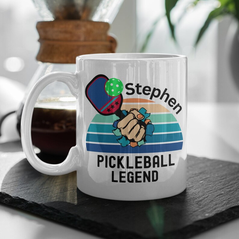 May include: White ceramic mug featuring a pickleball-themed design. The design includes a pickleball paddle, a green ball, and the text "Stephen PICKLEBALL LEGEND" against a retro sunset background. The mug has a curved handle.