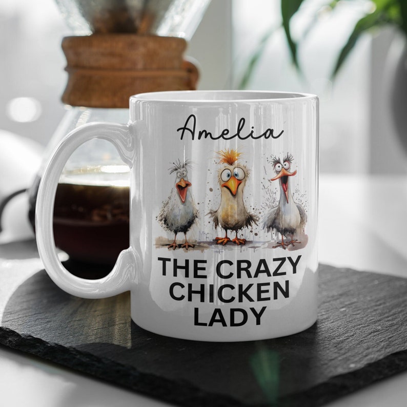 Puede incluir: Taza de cer&aacute;mica blanca con tres pollos de dibujos animados con expresiones divertidas. La taza tiene el nombre "Amelia" y el texto "THE CRAZY CHICKEN LADY". La taza est&aacute; sobre una superficie negra.