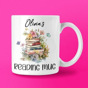 May include: White ceramic mug with the text "Olivia's reading mug". The mug features a watercolour illustration of a stack of books, flowers, and butterflies. The handle is white and the background is a bright pink.