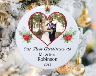 Op de afbeelding: Ronde witte keramische kerstversiering met een foto van een stel in een hartvormige lijst. De versiering heeft rode kralenranden, bloemmotieven en de tekst "Our First Christmas as Mr & Mrs Robinson 2023".