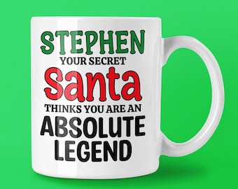 May include: White ceramic mug with the text "STEPHEN YOUR SECRET Santa THINKS YOU ARE AN ABSOLUTE LEGEND" in green, red, and black lettering. The mug has a curved handle and is set against a green background.
