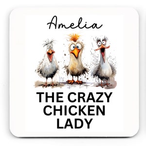 Puede incluir: Posavasos cuadrado blanco con una ilustraci&oacute;n de dibujos animados de tres pollos con expresiones exageradas. El nombre "Amelia" est&aacute; escrito encima de los pollos, y el texto "THE CRAZY CHICKEN LADY" est&aacute; debajo en negrita, en negro.