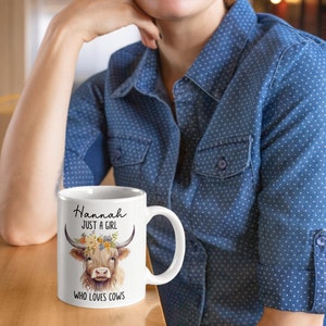 May include: White ceramic mug featuring a watercolor illustration of a Highland cow with floral accents and the text "Hannah JUST A GIRL WHO LOVES COWS". The mug has a curved handle and is set on a wooden surface.