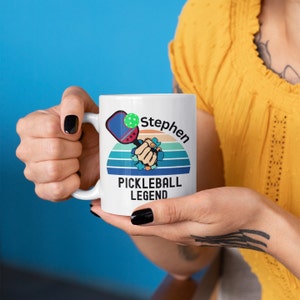 May include: White ceramic mug featuring a pickleball design with a paddle and ball, a fist breaking through a striped background, and the text "Stephen PICKLEBALL LEGEND". The mug is held by a person.