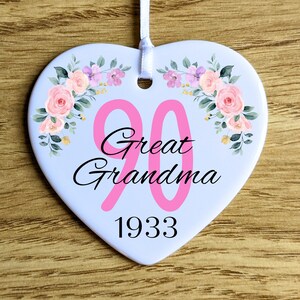 Può includere: Ornamento a forma di cuore con decorazioni floreali e la scritta "90 Great Grandma 1933". L'ornamento presenta rose rosa e fiori viola. Un nastro bianco è attaccato per appenderlo. Lo sfondo è in legno.