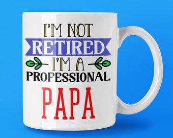 May include: White ceramic mug with the text "I'M NOT RETIRED I'M A PROFESSIONAL PAPA" in black, blue, green, and red lettering. The mug has a white handle and is set against a blue background. A gift for a father.