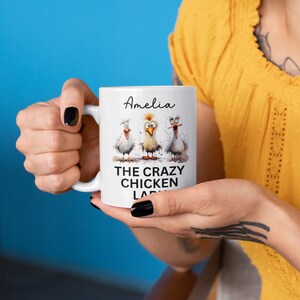 Puede incluir: Taza de cer&aacute;mica blanca con tres pollos de dibujos animados con expresiones divertidas. La taza tiene el texto "Amelia" arriba y "THE CRAZY CHICKEN LADY" abajo. La taza es sostenida por una persona con esmalte de u&ntilde;as negro.