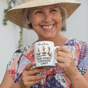 Puede incluir: Taza de cer&aacute;mica blanca con tres pollos de dibujos animados y el texto "THE CRAZY CHICKEN LADY" y el nombre "Amelia". La taza tiene un asa blanca y la sostiene una persona.