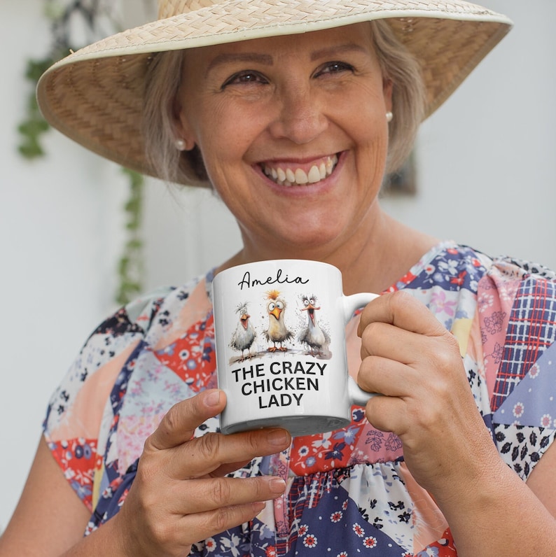 Puede incluir: Taza de cer&aacute;mica blanca con tres pollos de dibujos animados y el texto "THE CRAZY CHICKEN LADY" y el nombre "Amelia". La taza tiene un asa blanca y la sostiene una persona.