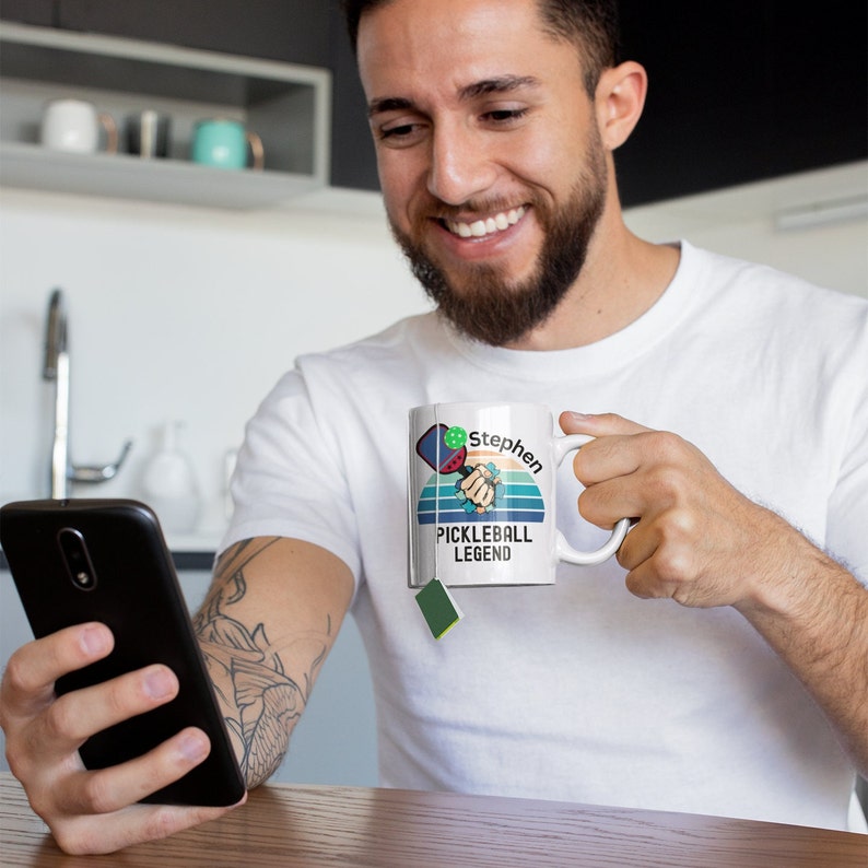 May include: White ceramic mug featuring a pickleball paddle and ball design with the text "Stephen PICKLEBALL LEGEND". The design includes a retro sunset and a hand holding the paddle. The mug has a handle and is being held by a person.