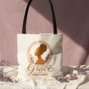 Può includere: Una borsa tote in tela color crema con spallacci neri. La parte anteriore presenta una grafica color oro di una silhouette di una persona con afro all'interno di una cornice ovale decorata, con il testo "Covered in Grace" e "2 CORINTHIANS 12:9".