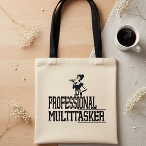 May include: Cream-colored tote bag with black handles featuring the text "PROFESSIONAL MULTITASKER" in bold, black letters. A black and white graphic of a woman holding a tray is above the text. A cup of coffee and white flowers are in the background.