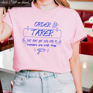 May include: Pink t-shirt with blue text that reads "ORDER TAKER but don't get cute with it manners are still free".