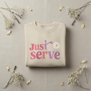 May include: A beige sweatshirt with the words "Just Serve" in pink and purple, with a daisy graphic. The sweatshirt is surrounded by sprigs of white flowers and petals on a neutral background.