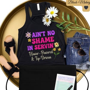 Könnte beinhalten: Schwarzes Tanktop mit dem Text "Ain't No Shame In Servin' Flower-Powered & Tip-Driven" in rosa und lila Schrift. Das Tanktop liegt auf einem goldenen Tablett mit einer schwarzen Schürze und einem Notizblock mit einem Stift.