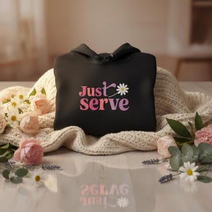 May include: Black hoodie with the text "Just Serve" in pink and white, with a daisy graphic. The hoodie is displayed on a cream-colored knitted blanket, surrounded by flowers.