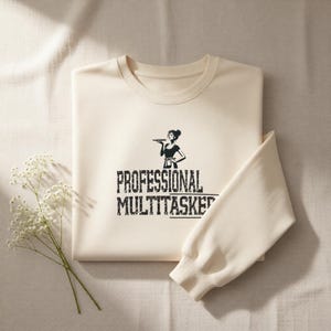 Puede incluir: Sudadera color crema con el texto negro "PROFESSIONAL MULTITASKER" y un gráfico de una mujer sosteniendo una bandeja. La manga de la sudadera está doblada y una ramita de flores blancas está en la esquina.