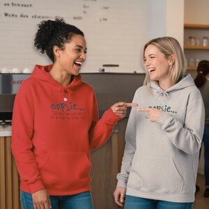 May include: Two people wearing matching hoodies. One is red, the other is gray. Both hoodies have the word "oopsie..." printed on the front, along with additional text. The hoodies have a front pocket and a drawstring hood.
