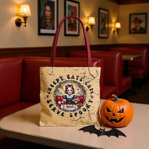 Puede incluir: Un bolso tote beige con asa burdeos, con una ilustración de estilo vintage de una chica en un café, con el texto "Creepy Eats Cafe Serve Spooky". También hay una calabaza naranja tallada y un murciélago negro.