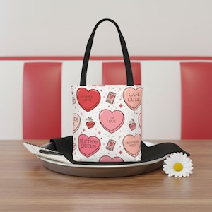 May include: White tote bag with black handles featuring a pattern of pink and red heart shapes with phrases like "Coffee Cutie" and "Tip Love." The bag is sitting on a metal tray on a wooden table.