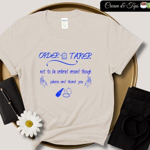 May include: A beige t-shirt with blue text that reads "ORDER TAKER not to be ordered around though please and thank you".