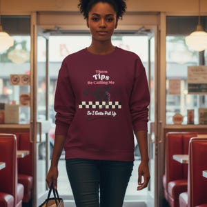 May include: Burgundy sweatshirt with the text "Them Tips Be Calling Me So I Gotta Pull Up" in white and black. The design includes a vintage telephone and a checkered pattern. The sweatshirt is worn by a person.