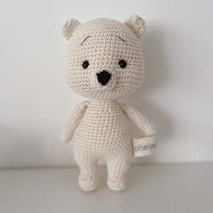 Könnte beinhalten: Ein gehäkelter cremefarbener Teddybär mit schwarzen Augen und einer schwarzen Nase. Der Bär steht auf einer weißen Oberfläche. Der Bär hat ein kleines Etikett am Arm, auf dem "ohhello" steht.