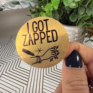 May include: Gold round sticker with the text "I got zapped" in black lettering. The sticker features a black and white illustration of a hand with a needle and a star.