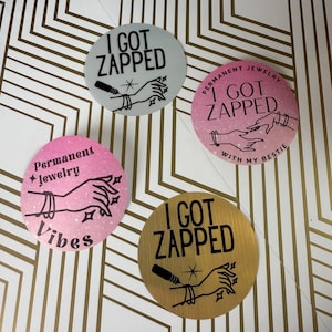 May include: Three round stickers with a black and white illustration of hands getting a tattoo. The stickers have different backgrounds: light blue, pink, and gold. The text on the stickers reads "I got zapped", "Permanent Jewelry Vibes", and "Permanent Jewelry I got zapped with my bestie".