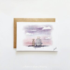 May include: Watercolor art print of a person and a dog sitting together, facing away from the viewer, with a purple and pink sky and clouds in the background. The art is on a white card with a brown envelope.