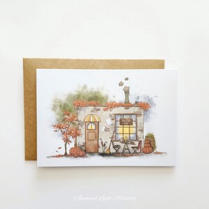 May include: Watercolor illustration of a cozy cafe in autumn. The building has a brown door and a window with the text "Big Girl Cafe". Pumpkins and fall foliage decorate the scene. The card is paired with a brown envelope.