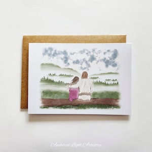 May include: A greeting card featuring an illustration of two figures seated on a log, overlooking a misty landscape. One figure wears a white robe, the other a pink dress. The card is set against a brown envelope.