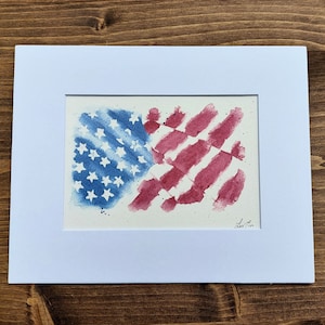 May include: A watercolor painting of an American flag with red and blue stripes and white stars. The flag is painted in a loose, abstract style. The painting is signed and dated "2021-12".