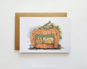 May include: A greeting card featuring an illustration of a pumpkin with a pastry shop built into it. The shop has a green awning and is lit by two lanterns. The card is on a brown envelope.