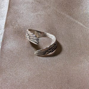 May include: A silver-colored ring with a feather design. The ring wraps around the finger with the feather detail on the top. The ring is on a tan fabric surface.