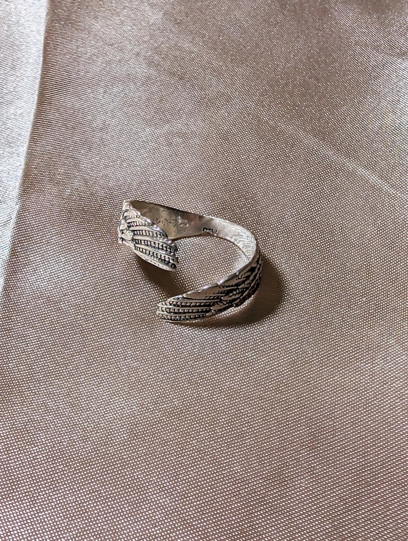 May include: A silver-colored ring with a feather design. The ring wraps around the finger with the feather detail on the top. The ring is on a tan fabric surface.