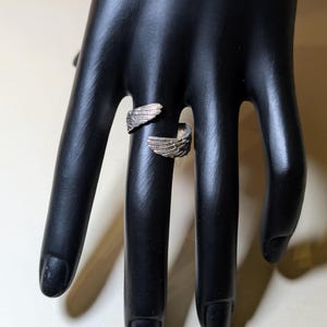 May include: A silver-toned ring with an open design, featuring wing-shaped details on each end. The ring is displayed on a black hand, showcasing its intricate design and metallic sheen. The wings are detailed with individual feather lines.