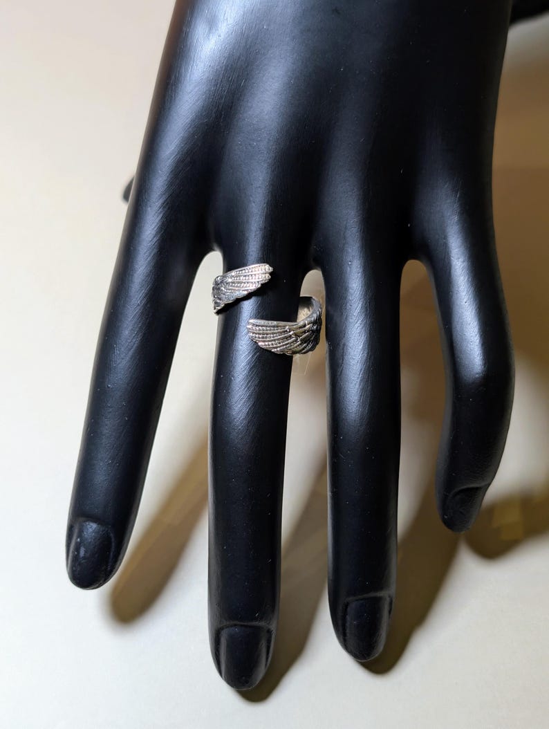 May include: A silver-toned ring with an open design, featuring wing-shaped details on each end. The ring is displayed on a black hand, showcasing its intricate design and metallic sheen. The wings are detailed with individual feather lines.