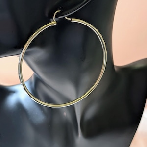 May include: A large, thin, circular hoop earring crafted from polished gold-tone metal. The earring features a smooth, continuous wire design with a simple hinged closure mechanism at the top, creating a classic and minimalist jewelry piece.