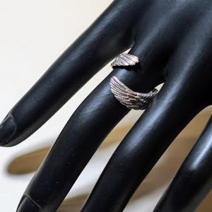 May include: A silver-colored ring with a wing design. The ring wraps around the finger, with the wings extending outwards. The ring is displayed on a black hand.