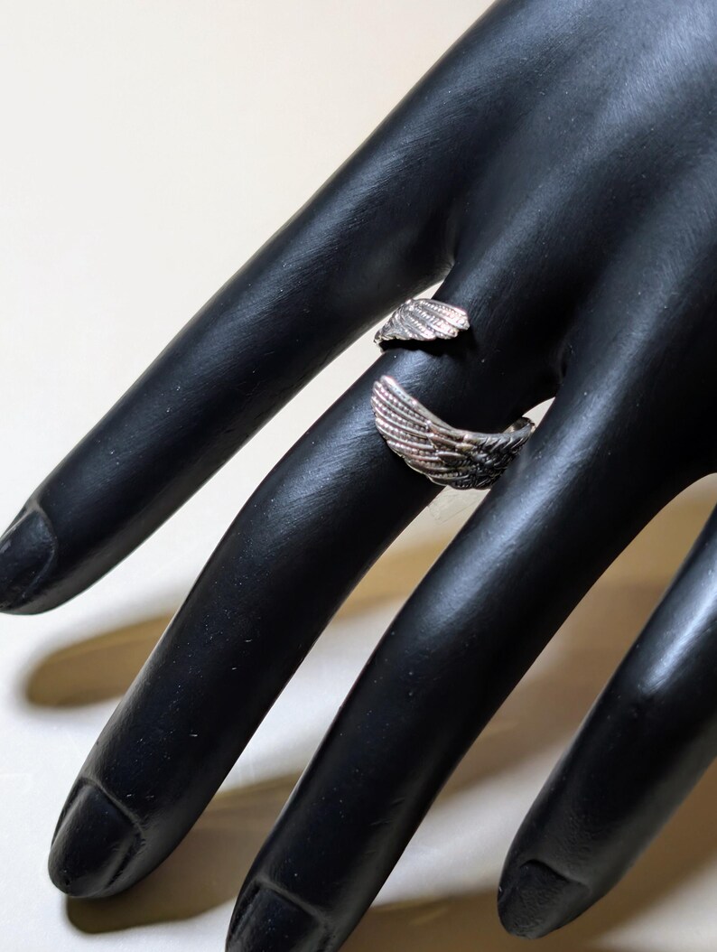 May include: A silver-colored ring with a wing design. The ring wraps around the finger, with the wings extending outwards. The ring is displayed on a black hand.
