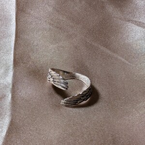 May include: A silver-colored ring with a detailed feather design. The ring is open, allowing for adjustable sizing. The feathers are intricately carved, adding texture and visual interest to the jewelry. The ring is set against a neutral background.