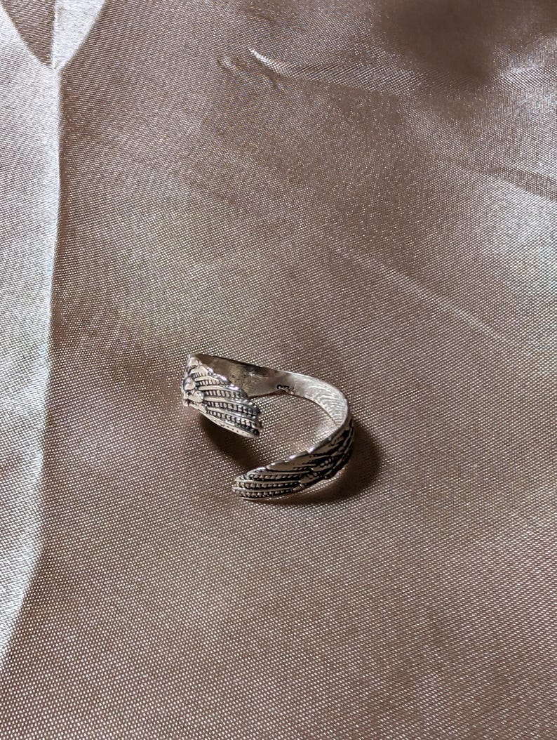 May include: A silver-colored ring with a detailed feather design. The ring is open, allowing for adjustable sizing. The feathers are intricately carved, adding texture and visual interest to the jewelry. The ring is set against a neutral background.
