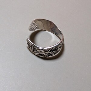 May include: A silver-colored ring with a feather design. The ring is open, with the feather pattern wrapping around the band. The ring is on a neutral gray surface.