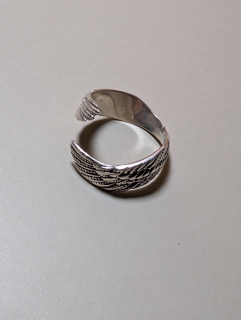 May include: A silver-colored ring with a feather design. The ring is open, with the feather pattern wrapping around the band. The ring is on a neutral gray surface.