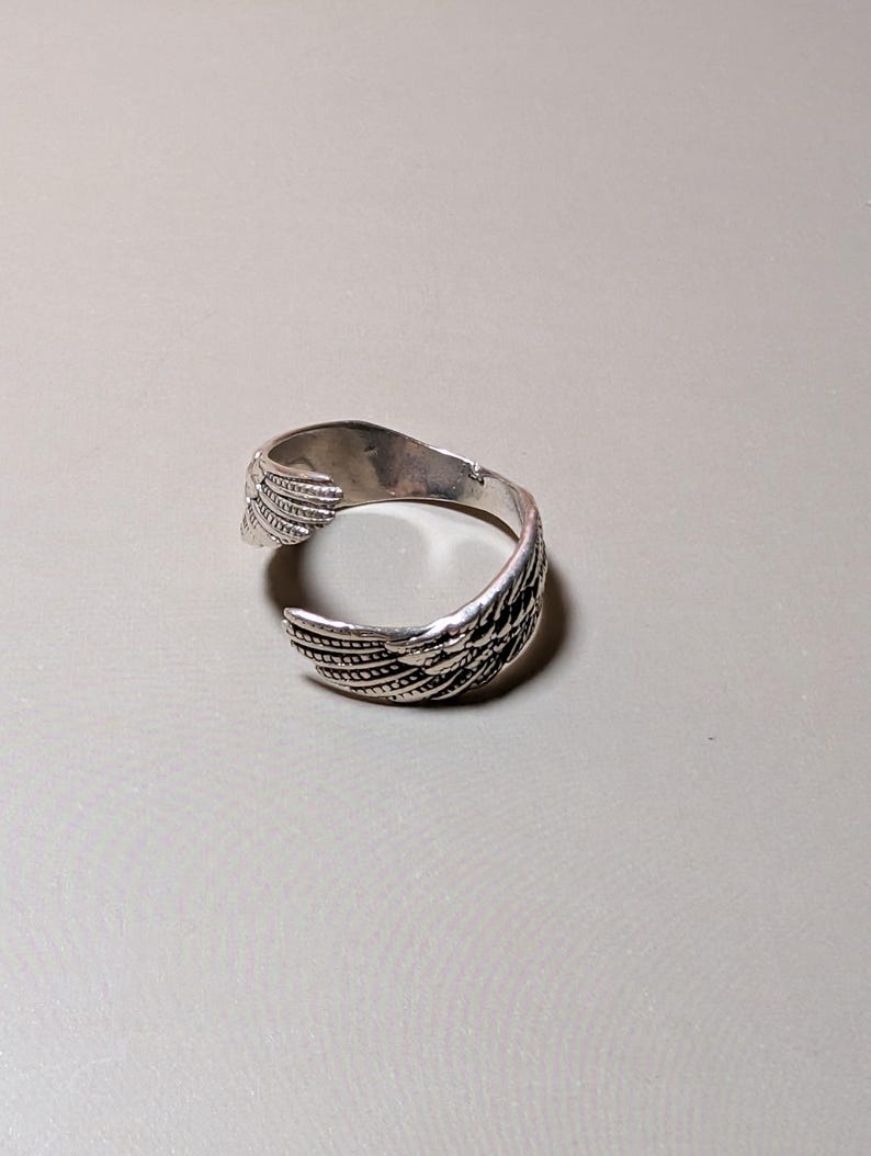 May include: A silver-toned cuff bracelet with a wing design. The open cuff features detailed feather patterns, creating a textured appearance. The bracelet is designed to wrap around the wrist.