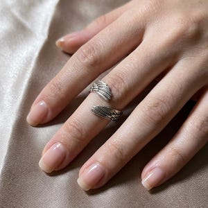 May include: Two silver rings, each featuring a detailed wing design, are worn on a hand. The rings are on the index and middle fingers. The nails are neatly manicured with a natural, glossy finish. The background is a soft, neutral color.